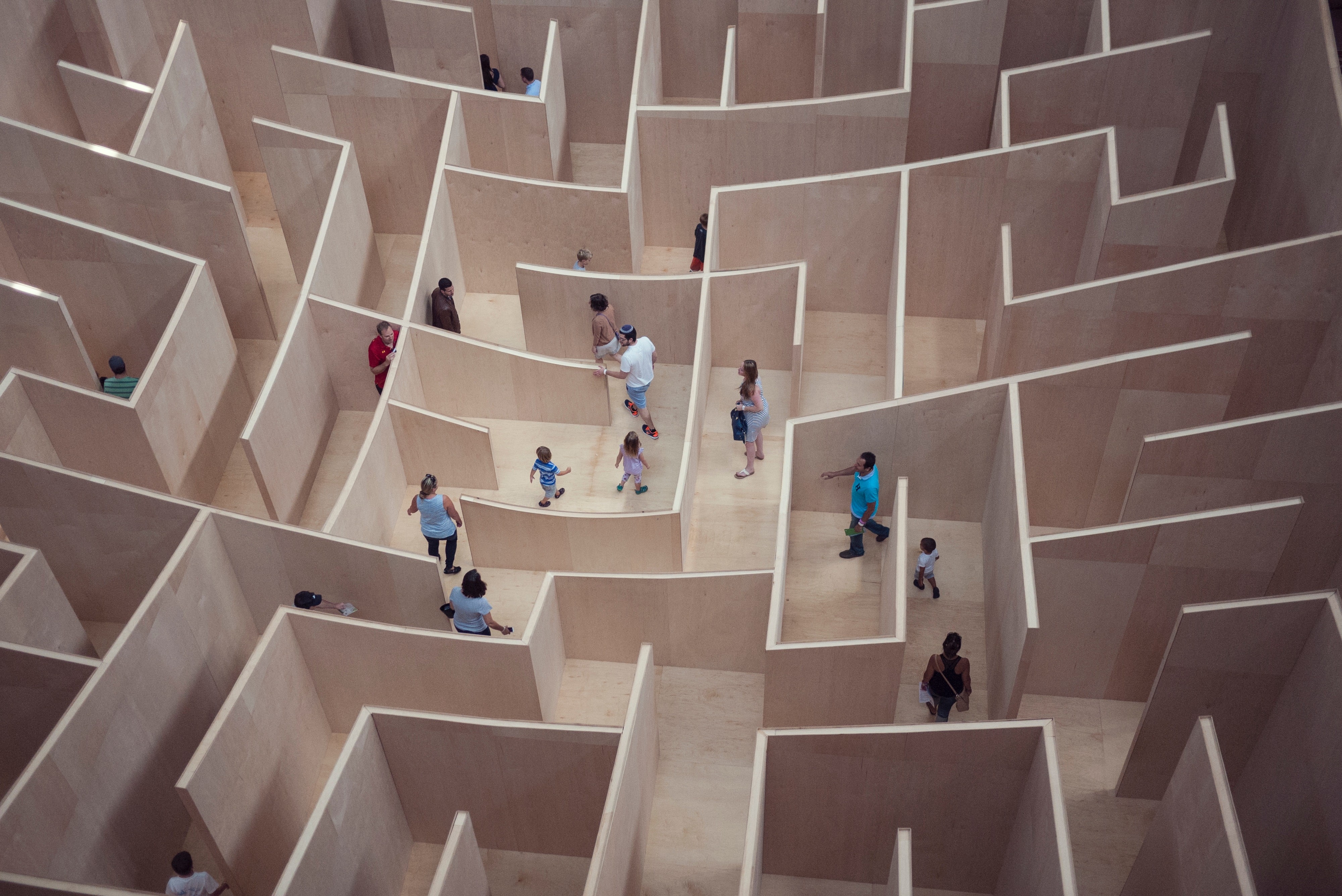 Auf dem Bild sind mehrere Menschen zu sehen, die durch ein Labyrinth gehen. Die Wände des Labyrinths sind dabei unterschiedlich hoch, ein Mann guckt sogar über eine der Wände.